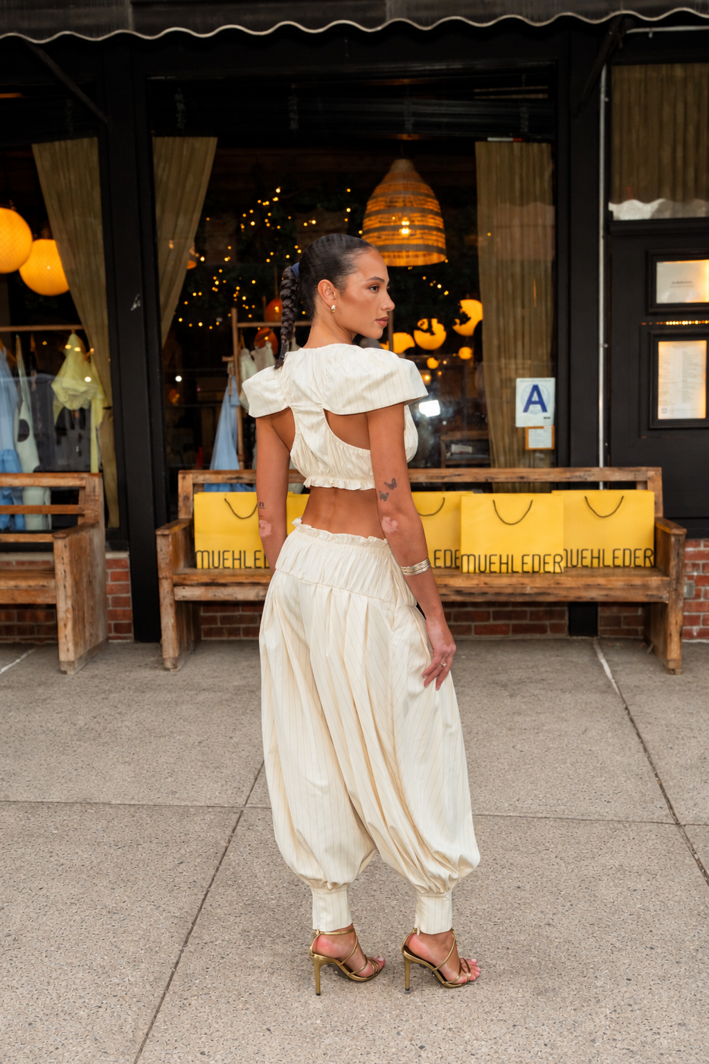 A woman stands on a sidewalk in front of a storefront, wearing a cream-colored two-piece outfit consisting of a ruffled puff-sleeve crop top and matching high-waisted balloon harem pants with elastic ankle cuffs.