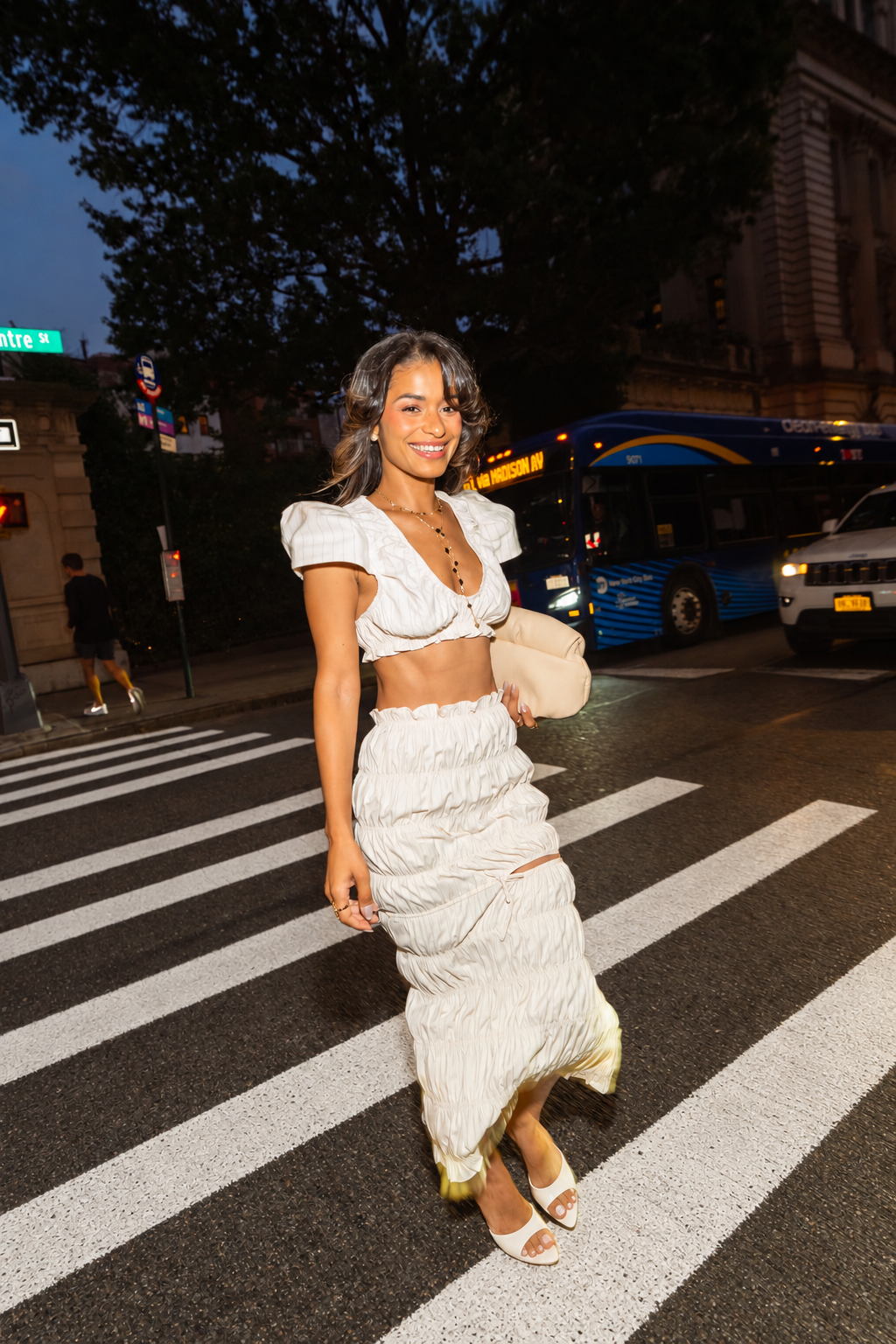 A woman walking across a city crosswalk at dusk wearing a white two-piece outfit consisting of a short puff-sleeve crop top and a matching pleated maxi skirt, holding a beige clutch.