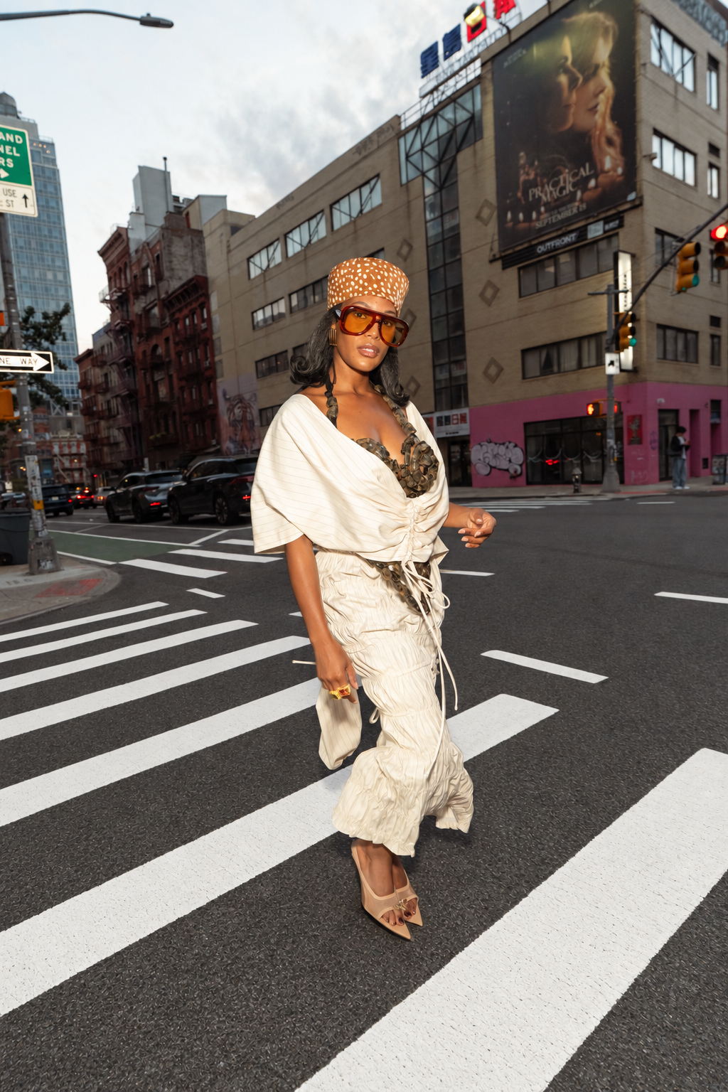 A woman walking across a city crosswalk wearing a cream-colored draped wrap top and matching wide-leg pants, a patterned orange hat, and sunglasses, with a busy urban street background.