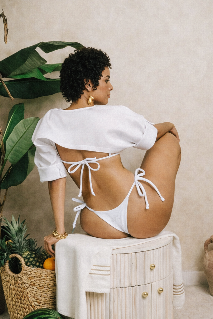 Woman in a white bikini sitting on a stool with plants around her.
