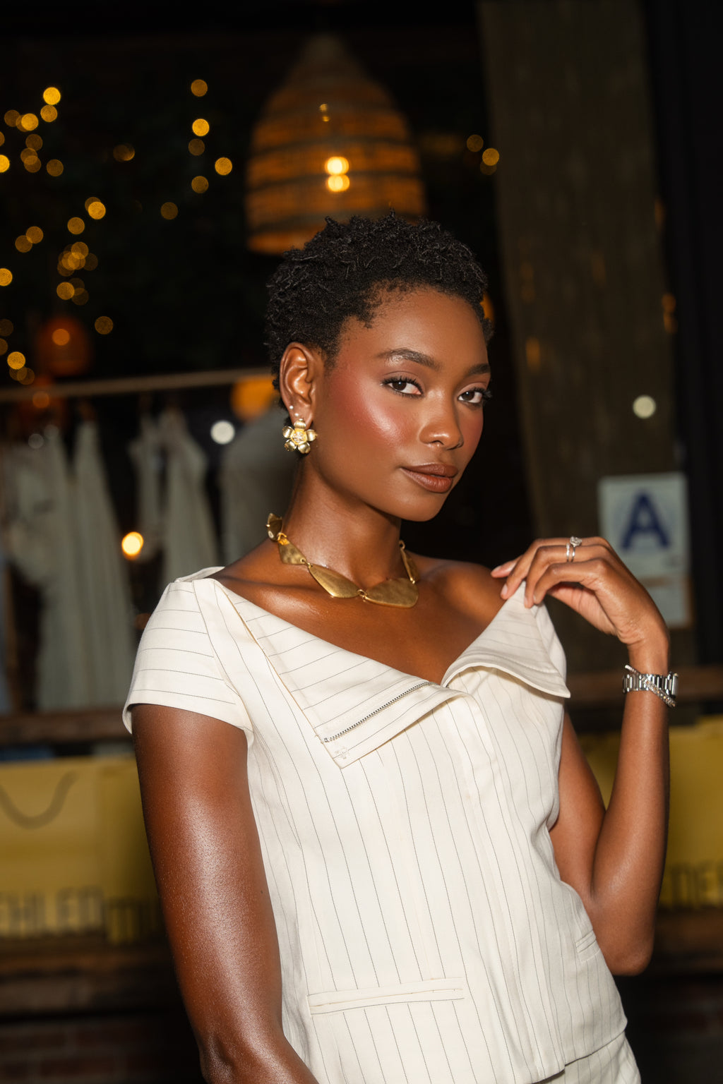 A woman with short dark hair wearing a cream-colored pinstriped top and gold jewelry, posing in a dimly lit, warm-toned indoor setting.