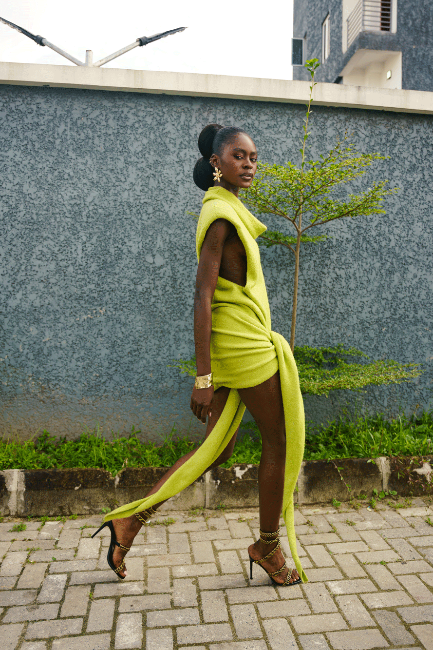 Woman in a bright green dress walking on a sidewalk with a building and tree in the background.